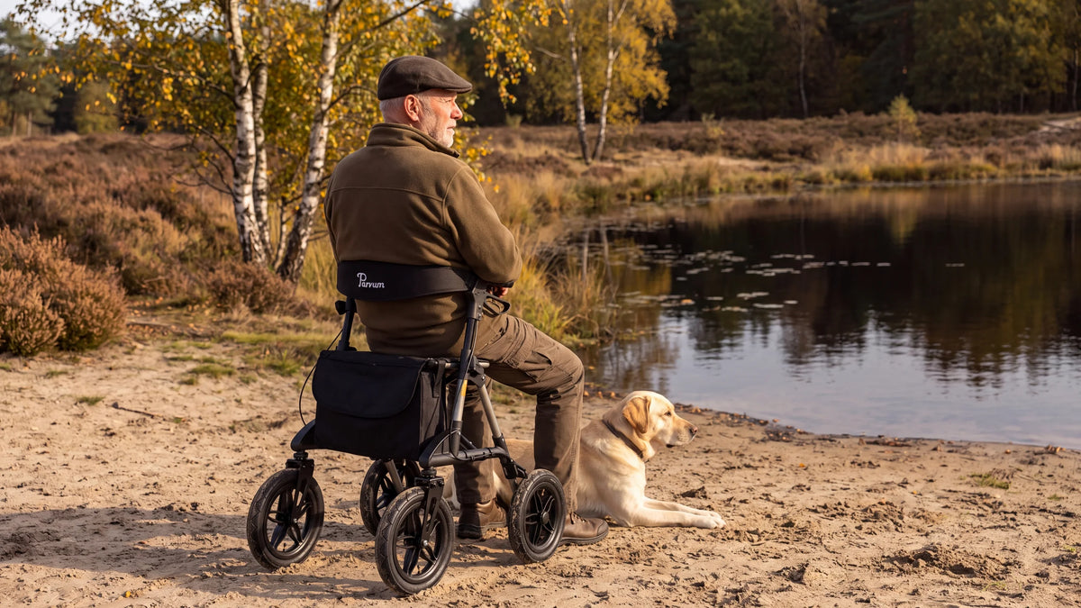 Parvum Rebel in gray by a heathland pond; a man sits on the rollator, his Labrador lying beside him.